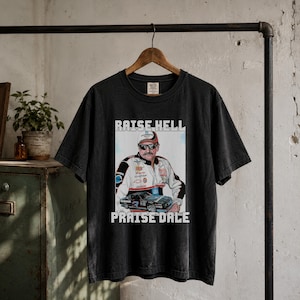 May include: A black short-sleeve t-shirt featuring a graphic print of a race car driver wearing a cap and sunglasses, standing in front of a race car. The text RAISE HELL is printed above the image and PRAISE DALE is printed below the image.
