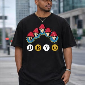 May include: Black t-shirt featuring a graphic of five stylized faces wearing red hats, surrounded by a blue and yellow design. The word "DEVO" is printed below in white and yellow circles.