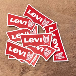 May include: A collection of red and white Levi's logo stickers. Each sticker features the Levi's name in white lettering on a red background, with a white leaf design. The stickers are scattered on a brown surface.
