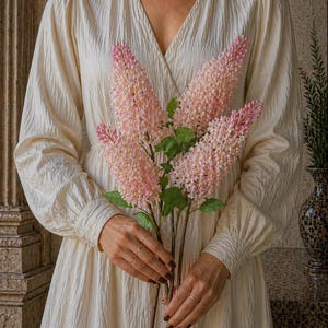 Puede incluir: Una persona sostiene un ramo de flores rosas y blancas con hojas verdes. Las flores son largas y delgadas, con una apariencia texturizada. La persona lleva un vestido color crema con mangas largas y cuello en V.