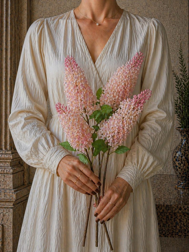 Puede incluir: Una persona sostiene un ramo de flores rosas y blancas con hojas verdes. Las flores son largas y delgadas, con una apariencia texturizada. La persona lleva un vestido color crema con mangas largas y cuello en V.