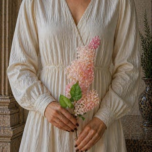Puede incluir: Un vestido color crema con cuello en V y mangas largas. El vestido tiene una tela texturizada y una cintura fruncida. Se sostiene una ramita de flores rosas frente al vestido. El vestido es de estilo cl&aacute;sico y elegante.
