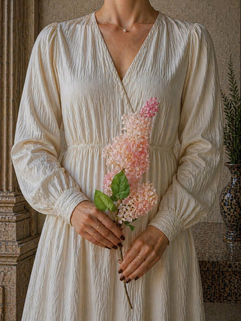 Puede incluir: Un vestido color crema con cuello en V y mangas largas. El vestido tiene una tela texturizada y una cintura fruncida. Se sostiene una ramita de flores rosas frente al vestido. El vestido es de estilo cl&aacute;sico y elegante.