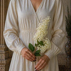 Puede incluir: Vestido crema de manga larga con cuello en V y tejido texturizado. El vestido presenta una cintura fruncida y es sostenido por una persona que sostiene una ramita de flores blancas. La persona lleva anillos dorados y un delicado collar.