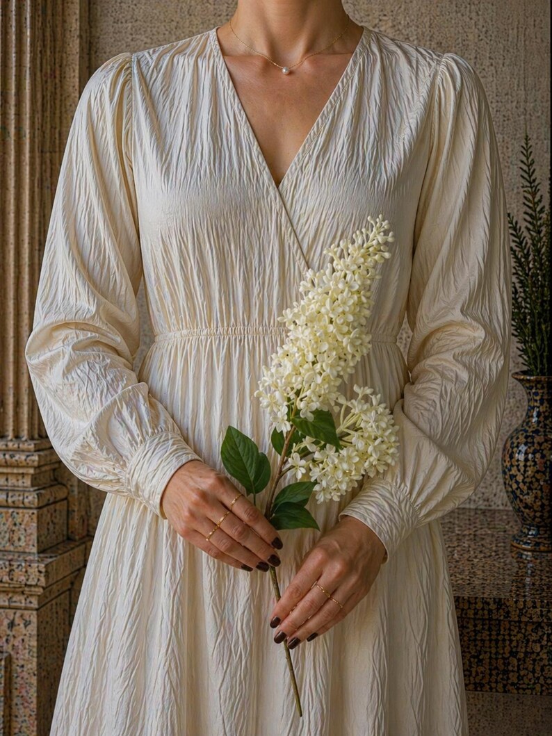 Puede incluir: Vestido crema de manga larga con cuello en V y tejido texturizado. El vestido presenta una cintura fruncida y es sostenido por una persona que sostiene una ramita de flores blancas. La persona lleva anillos dorados y un delicado collar.