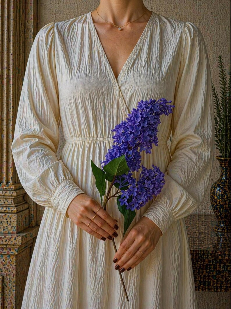 Puede incluir: Vestido crema de manga larga con cuello en V y tejido texturizado. Una persona sostiene el vestido y una ramita de flores moradas. El vestido tiene cintura entallada y falda fluida.