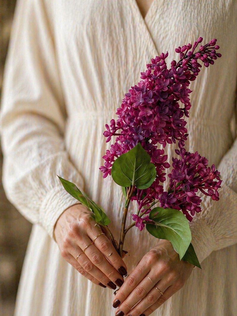 Puede incluir: Una rama de lilas moradas vibrantes con hojas verdes. Las flores se sostienen en manos con anillos dorados, contra un vestido color crema. La imagen resalta la belleza natural de las flores.