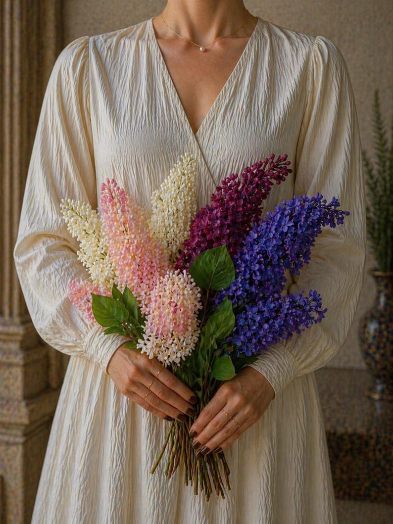 Puede incluir: Un ramo de flores de lilas coloridas en tonos blancos, rosas, morados y azules. Las flores se sujetan con hojas verdes y tallos marrones. La persona lleva un vestido color crema.