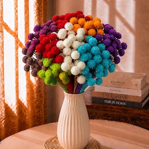 Puede incluir: Un ramo de flores secas billy button en tonos vibrantes de rojo, naranja, amarillo, blanco, turquesa, morado, verde lima y marrón, dispuesto en un jarrón de cerámica alto de color blanco roto con nervaduras verticales, cuello estrecho y base.