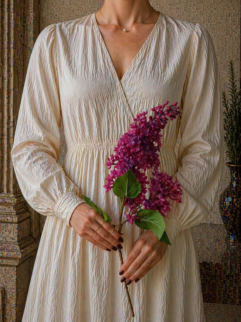 Puede incluir: Vestido crema de manga larga con cuello en V y tejido texturizado. El vestido est&aacute; recogido en la cintura y sostenido por una persona que sostiene una ramita de lilas moradas. Se ve un collar de oro con un colgante de perla.