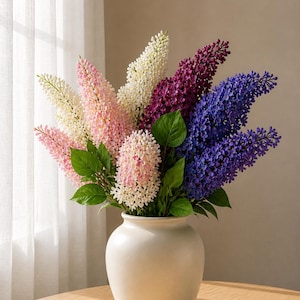 Puede incluir: Un ramo de lilas artificiales coloridas en un jarr&oacute;n de cer&aacute;mica color crema. Las flores son en tonos rosa, blanco, morado y azul, con hojas verdes. El jarr&oacute;n est&aacute; sobre una mesa de madera cerca de una ventana con cortinas transparentes.