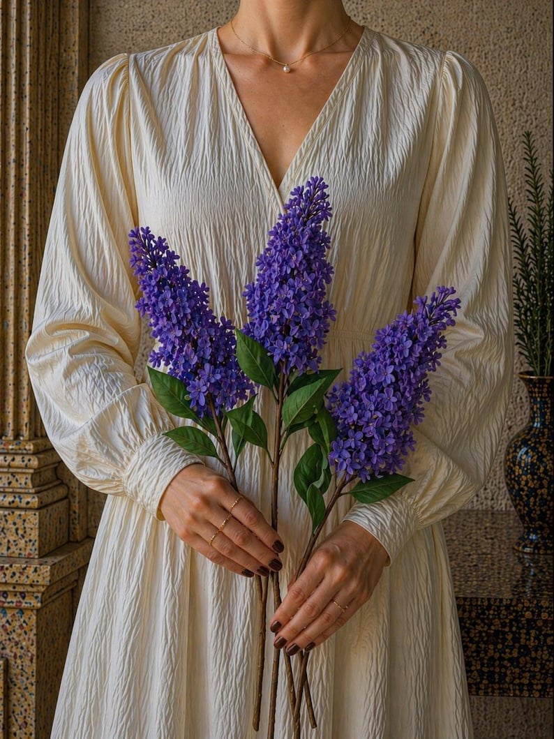 Puede incluir: Una persona sostiene tres tallos de flores de lilas moradas. Lleva un vestido color crema con mangas largas y cuello en V. El vestido tiene un patr&oacute;n texturizado. El fondo incluye un jarr&oacute;n decorativo y detalles arquitect&oacute;nicos.