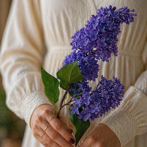 Puede incluir: Primer plano de una rama de vibrantes flores de lilas moradas con hojas verdes. Las flores se sostienen frente a un vestido color crema. La imagen resalta los detalles de las flores y la textura de la tela.