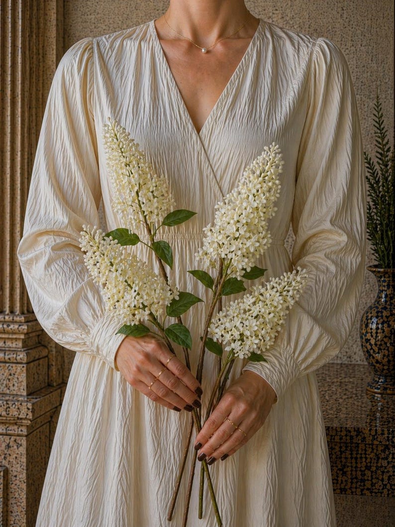 Puede incluir: Un vestido color crema con mangas largas y cuello en V. El vestido es texturizado y lo sostiene una persona. La persona sostiene tres tallos de flores blancas con hojas verdes. El fondo es de color neutro.