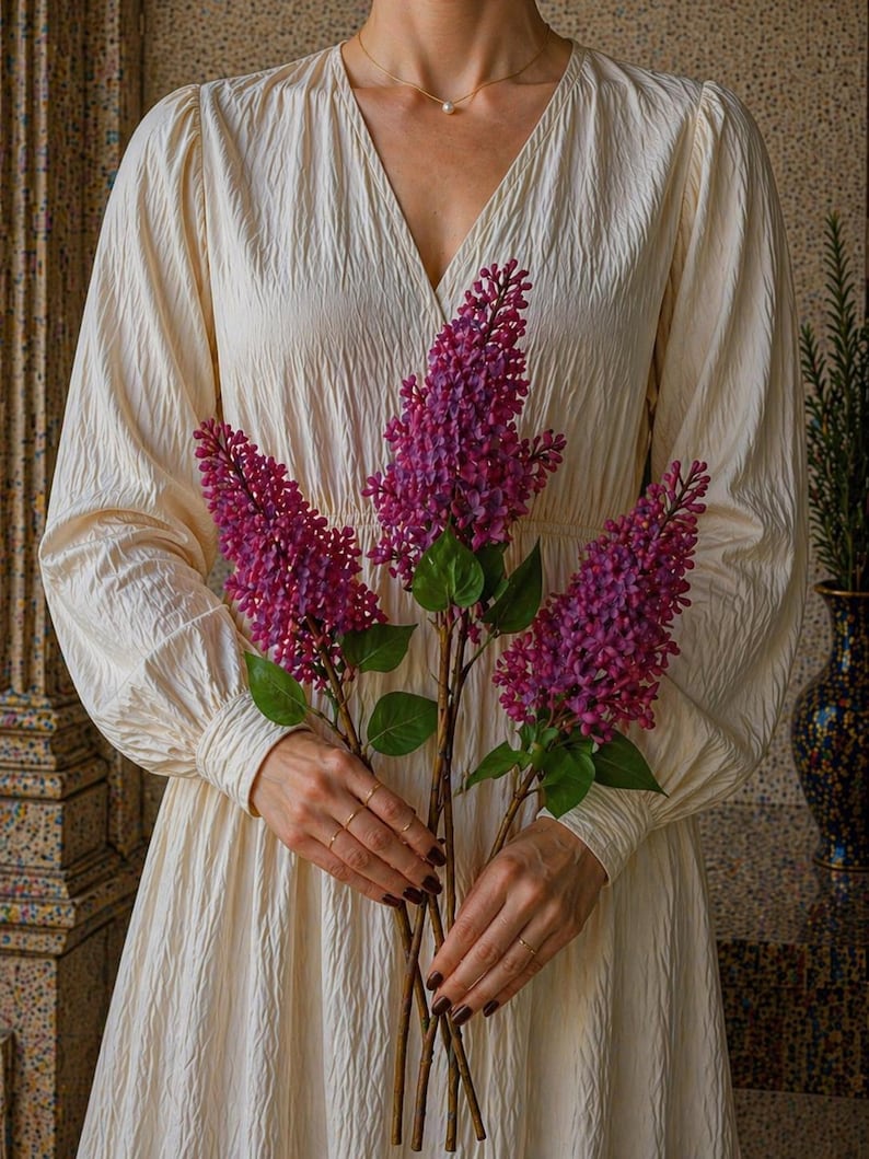 Puede incluir: Una persona con un vestido color crema con cuello en V sostiene tres tallos de lilas moradas. El vestido tiene mangas largas y una tela texturizada. Las flores tienen hojas verdes y est&aacute;n en plena floraci&oacute;n.