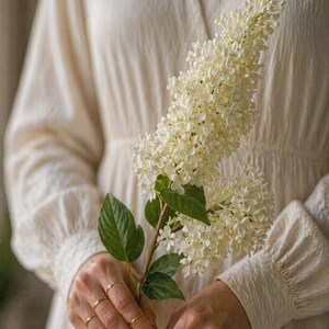 Puede incluir: Una persona con un vestido color crema con cuello en V y mangas largas sostiene una rama de flores blancas con hojas verdes. Varios anillos dorados adornan los dedos.