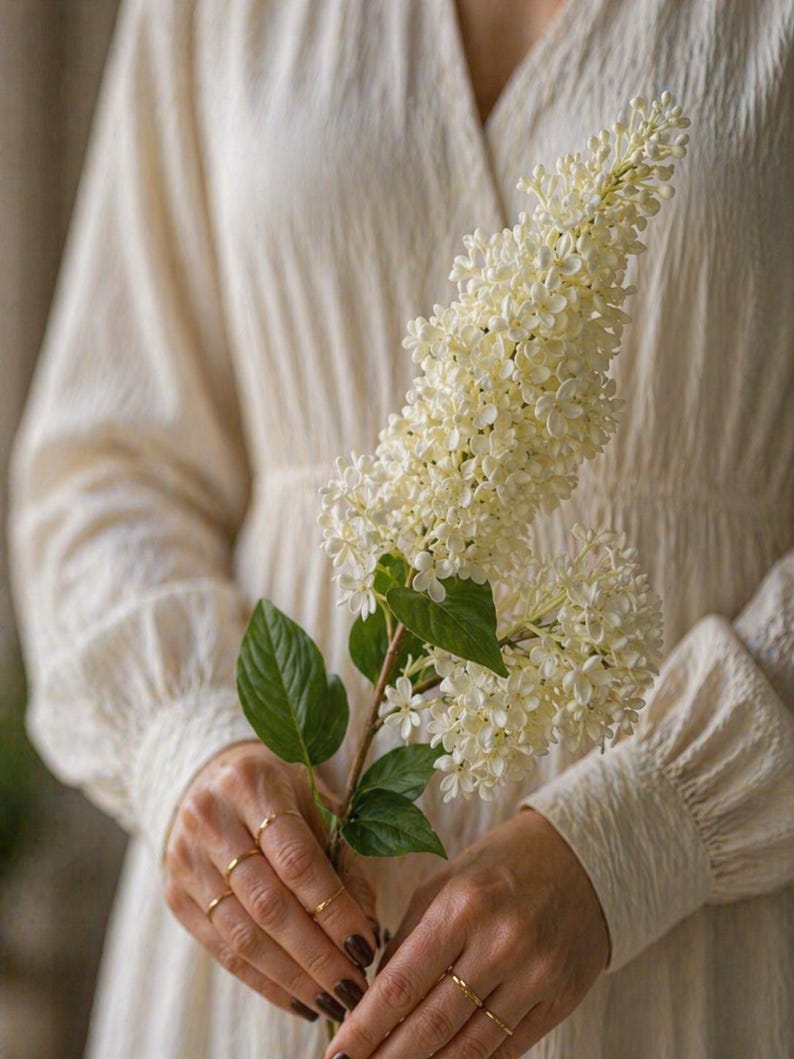 Puede incluir: Una persona con un vestido color crema con cuello en V y mangas largas sostiene una rama de flores blancas con hojas verdes. Varios anillos dorados adornan los dedos.