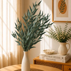 May include: A tall, slender, off-white textured vase holds a large arrangement of dark green eucalyptus-like branches with small berries. The vase sits on a round, woven brown placemat, creating a natural and simple decorative display.