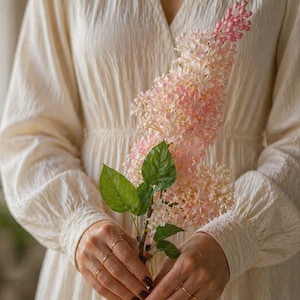 Puede incluir: Un vestido color crema con mangas largas y cuello en V. Las manos de una persona, adornadas con anillos dorados, sostienen un tallo de flores rosas y blancas con hojas verdes. Las flores tienen una apariencia delicada y en capas. El vestido tiene una tela texturizada.