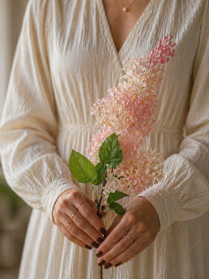 Puede incluir: Un vestido color crema con mangas largas y cuello en V. Las manos de una persona, adornadas con anillos dorados, sostienen un tallo de flores rosas y blancas con hojas verdes. Las flores tienen una apariencia delicada y en capas. El vestido tiene una tela texturizada.
