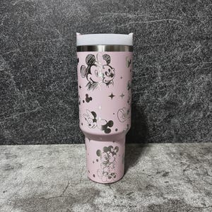 May include: A pink insulated tumbler featuring a stainless steel rim and white lid. The tumbler is adorned with silver illustrations of Mickey and Minnie Mouse, stars, and polka dots. The tumbler is designed to hold approximately 30 ounces.