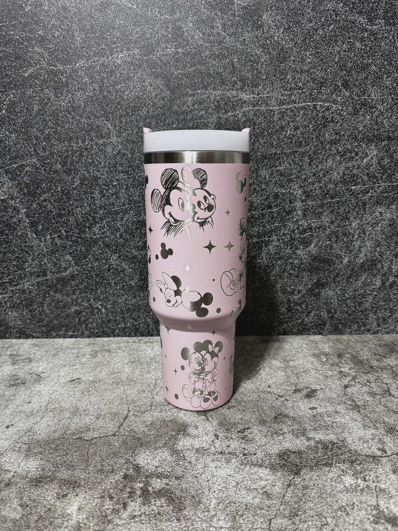 May include: A pink insulated tumbler featuring a stainless steel rim and white lid. The tumbler is adorned with silver illustrations of Mickey and Minnie Mouse, stars, and polka dots. The tumbler is designed to hold approximately 30 ounces.