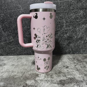 May include: A pink insulated tumbler with a handle, featuring a lid and a silver interior. The tumbler is decorated with silver illustrations of Mickey and Minnie Mouse, stars, and other shapes. The handle and lid accents are pink.