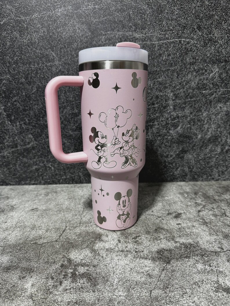 May include: A pink insulated tumbler with a handle, featuring a lid and a silver interior. The tumbler is decorated with silver illustrations of Mickey and Minnie Mouse, stars, and other shapes. The handle and lid accents are pink.