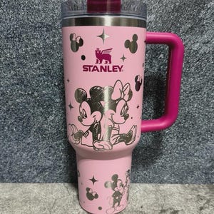 May include: Pink Stanley tumbler featuring Mickey and Minnie Mouse illustrations. The tumbler has a pink handle and lid, with the Stanley logo and star accents. The design includes a clear top and a stainless steel interior.