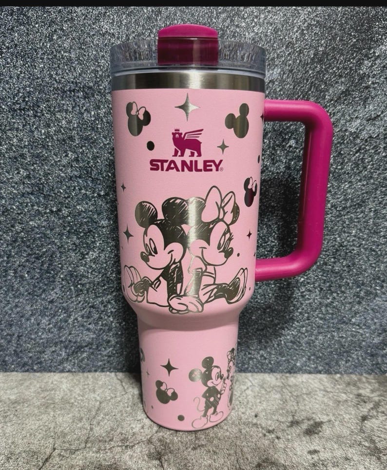 May include: Pink Stanley tumbler featuring Mickey and Minnie Mouse illustrations. The tumbler has a pink handle and lid, with the Stanley logo and star accents. The design includes a clear top and a stainless steel interior.