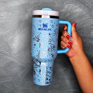 May include: A light blue Stanley travel mug with a white lid and a blue handle. The mug features a mermaid-themed design with black and white illustrations. The Stanley logo is visible on the front.