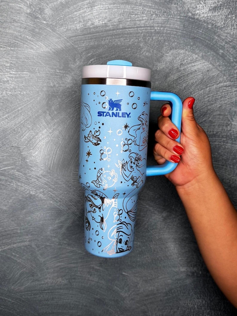 May include: A light blue Stanley travel mug with a white lid and a blue handle. The mug features a mermaid-themed design with black and white illustrations. The Stanley logo is visible on the front.