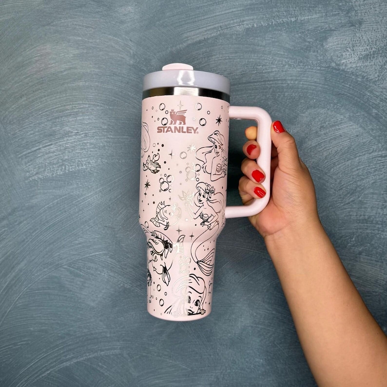 May include: A light pink Stanley travel mug with a handle, featuring black line art of mermaids and other sea creatures. The mug has a light gray lid and handle, and the Stanley logo is visible.