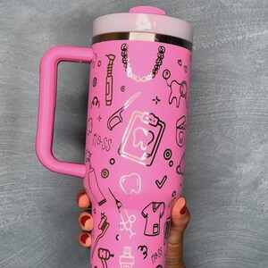 以下が含まれることがあります： ピンク色の断熱タンブラー。ハンドルと白い蓋が付いています。タンブラーには、歯や歯科用具など、歯科をテーマにしたイラストが描かれています。「Floss」の文字も確認できます。ハンドルと蓋は薄いピンク色です。