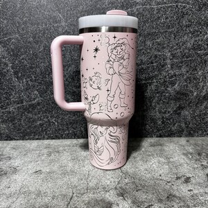 May include: A pink travel mug with a handle, featuring a stainless steel rim and lid. The mug is decorated with black line art of a princess and other characters. The handle and lid button are also pink.