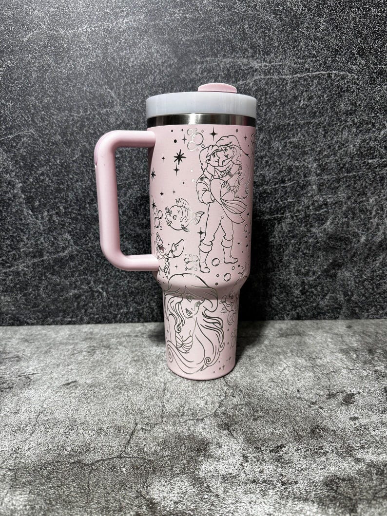 May include: A pink travel mug with a handle, featuring a stainless steel rim and lid. The mug is decorated with black line art of a princess and other characters. The handle and lid button are also pink.