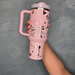 May include: Pink insulated tumbler with a handle, featuring silver illustrations of Mickey and Minnie Mouse, stars, and clouds. The lid and handle are also pink, with a silver rim. A fun and functional drinkware item.