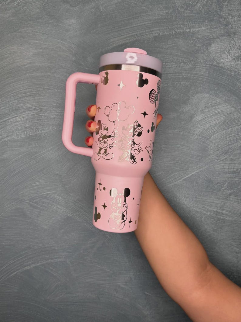 May include: Pink insulated tumbler with a handle, featuring silver illustrations of Mickey and Minnie Mouse, stars, and clouds. The lid and handle are also pink, with a silver rim. A fun and functional drinkware item.