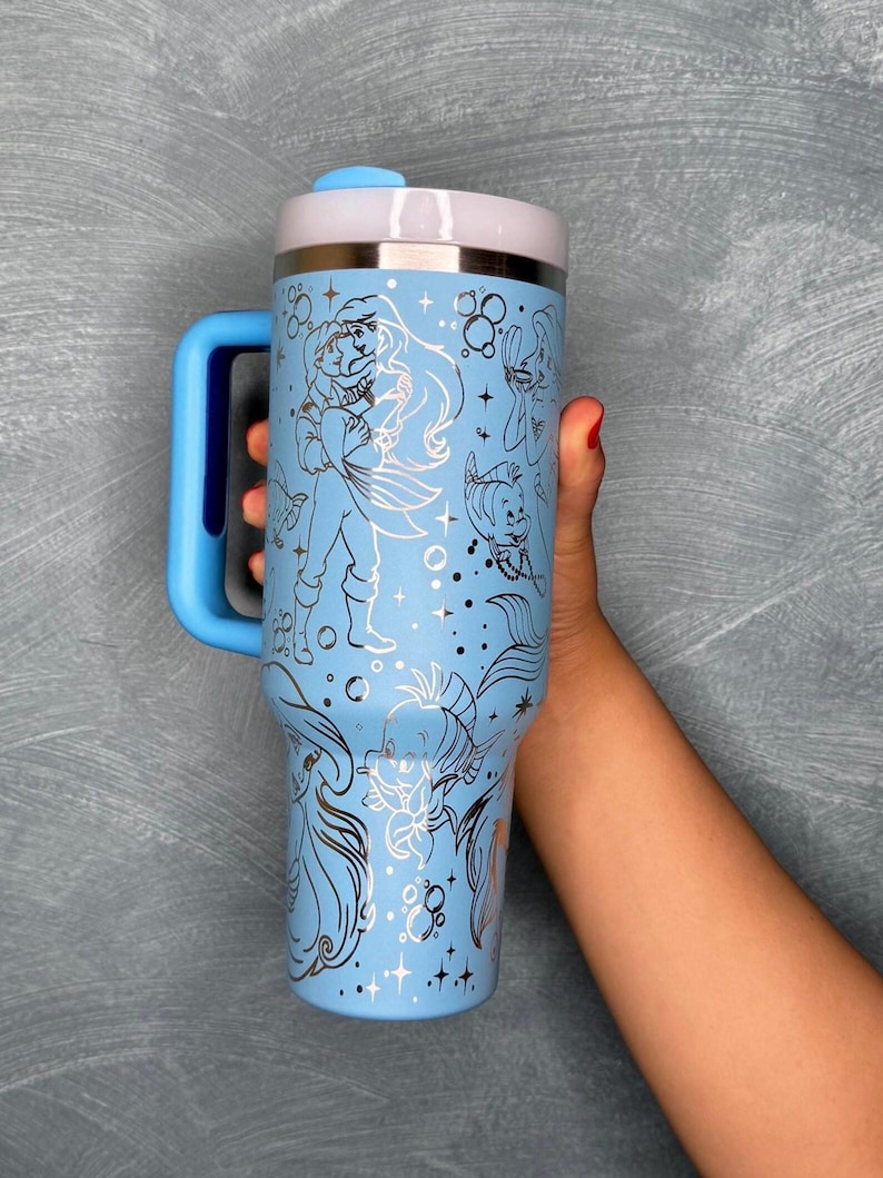 May include: A light blue insulated tumbler with a silver rim and a blue handle. The tumbler features line art illustrations of characters and elements from a popular animated film, including a mermaid, fish, and bubbles. The tumbler is held by a person.