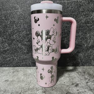 May include: A pink travel mug featuring Mickey and Minnie Mouse. The mug has a silver lid and handle, with silver Mickey Mouse silhouettes and stars. The design includes a large image of Mickey and Minnie Mouse. The mug is designed for hot or cold beverages.