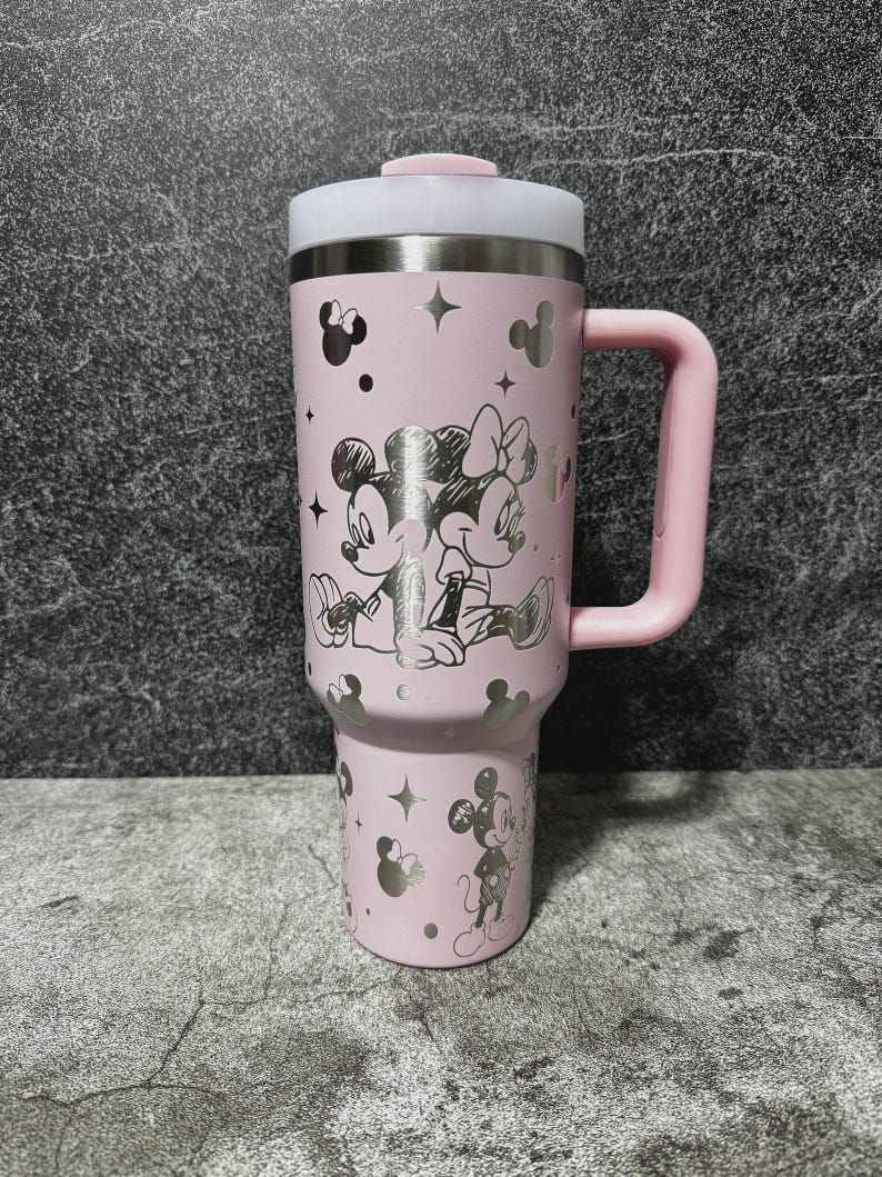 May include: A pink travel mug featuring Mickey and Minnie Mouse. The mug has a silver lid and handle, with silver Mickey Mouse silhouettes and stars. The design includes a large image of Mickey and Minnie Mouse. The mug is designed for hot or cold beverages.