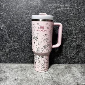 May include: A pink Stanley tumbler with a white lid and handle. The tumbler features a silver illustration of Disney princesses and the Stanley logo. The tumbler is designed for cold beverages and has a capacity of approximately 30 ounces.