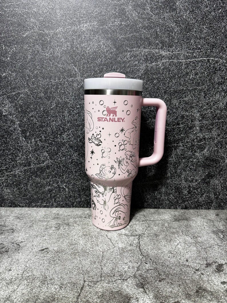 May include: A pink Stanley tumbler with a white lid and handle. The tumbler features a silver illustration of Disney princesses and the Stanley logo. The tumbler is designed for cold beverages and has a capacity of approximately 30 ounces.