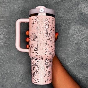May include: A pink insulated tumbler with a handle, featuring illustrations of characters from a popular animated film. The tumbler has a light pink lid and handle, and the design includes stars and other decorative elements.