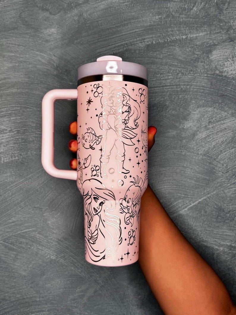 May include: A pink insulated tumbler with a handle, featuring illustrations of characters from a popular animated film. The tumbler has a light pink lid and handle, and the design includes stars and other decorative elements.