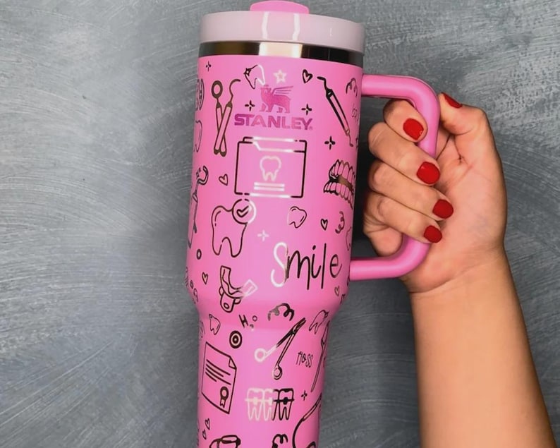 以下が含まれることがあります： ピンク色のStanleyタンブラー。歯科をテーマにしたイラストと「Smile」の文字が描かれています。タンブラーにはシルバーの縁と蓋があり、赤いマニキュアを塗った手で持っています。