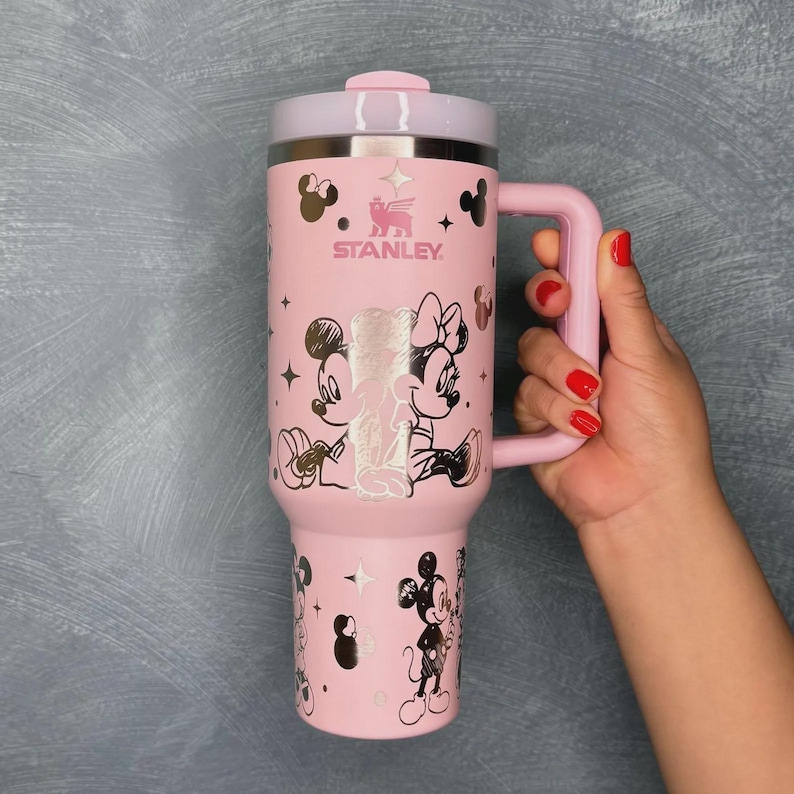 May include: Pink Stanley tumbler featuring Mickey and Minnie Mouse illustrations. The tumbler has a handle, a lid, and the Stanley logo. The design includes stars and character silhouettes. The tumbler is held by a person with red nail polish.