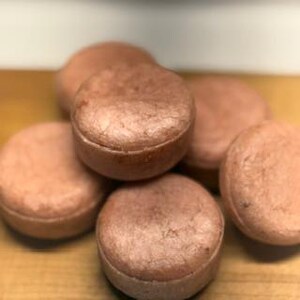 May include: A close-up of several round, light brown shampoo bars. The bars are stacked on top of each other, with some in the foreground and others in the background. The bars have a slightly textured surface and appear to be handmade.