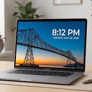May include: A silver MacBook Pro laptop displaying a bridge scene at sunset, with the time 8:12 PM and the date Tuesday, May 26, 2026. A white mug with "Infinite Studio" text and a pen are on the desk.