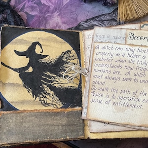 May include: An open book with a hand-drawn illustration of a witch flying on a broomstick against a large yellow moon. The adjacent page features handwritten text that reads: Becoming. A witch can only function properly as a healer & protector.
